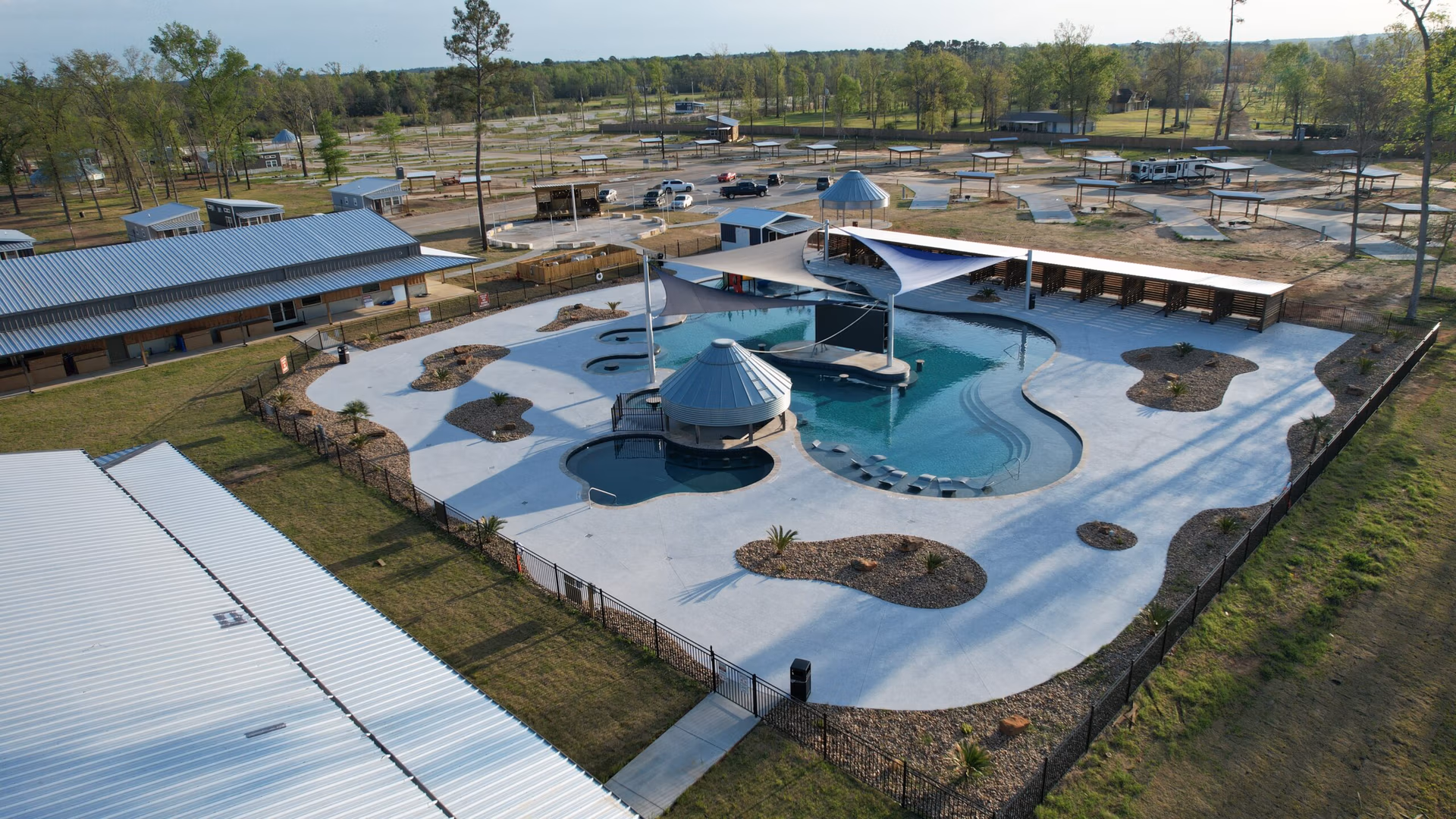 Aerial view of a modern swimming pool complex with a unique design. The pool is surrounded by a concrete patio with landscaped rock and plant areas. Canopies provide shade over parts of the pool. Buildings and open land are visible in the background.