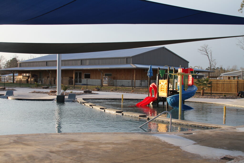 A large outdoor pool with colorful slides and shades over the water. A modern building is in the background, and the pool area is surrounded by a fence. Sparse greenery is visible around the perimeter.