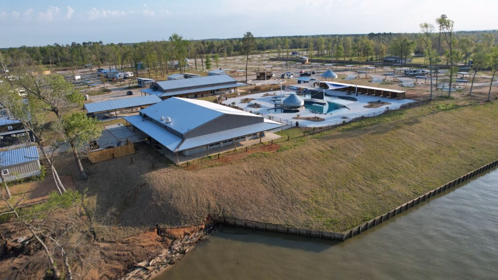 Aerial view of a resort with several modern buildings and a landscaped swimming pool area. The resort is near a wooded area and a body of water, with a grass embankment leading down to the water's edge.