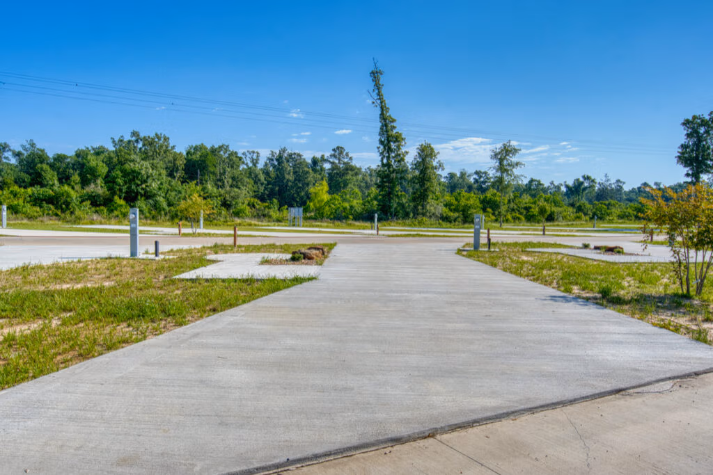 A wide, empty concrete lot with several utility hookups, surrounded by grass and trees under a clear blue sky. The area appears to be part of an RV park or campground.