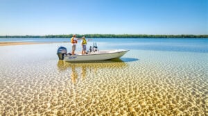 Two anglers in a small boat near exposed sandbar on a calm lake, with distant trees and clear water, under bright midday sunlight.