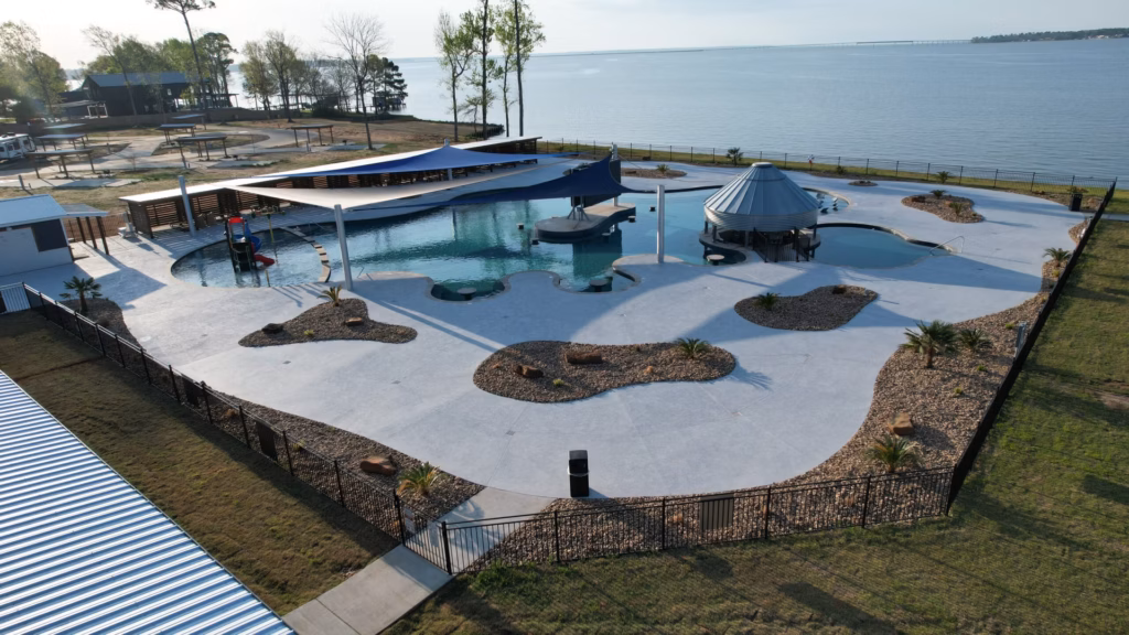 Aerial view of a lakeside outdoor pool area with shaded seating, small islands of landscaping, adjacent to a large body of water and surrounded by a metal fence.