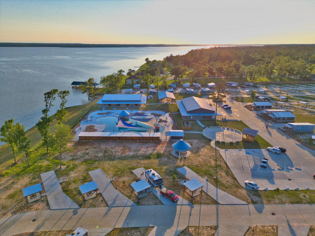 Aerial view of a lakeside recreation area with a pool, parking lots, picnic shelters, RVs, and nearby forested shoreline at sunset.