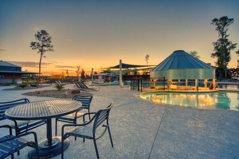 Outdoor pool area at sunset with lounge chairs, tables, a covered pool section, and shaded play structures visible in the background.
