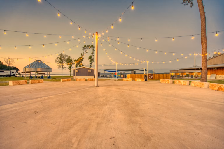 An outdoor venue with string lights overhead, stone benches arranged in a circle, and buildings and trees in the background at sunset.