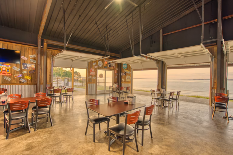Open-air restaurant with red and black chairs, wooden walls, and large garage doors raised to reveal a waterfront view at sunset. Tables are spaced apart on a polished concrete floor.