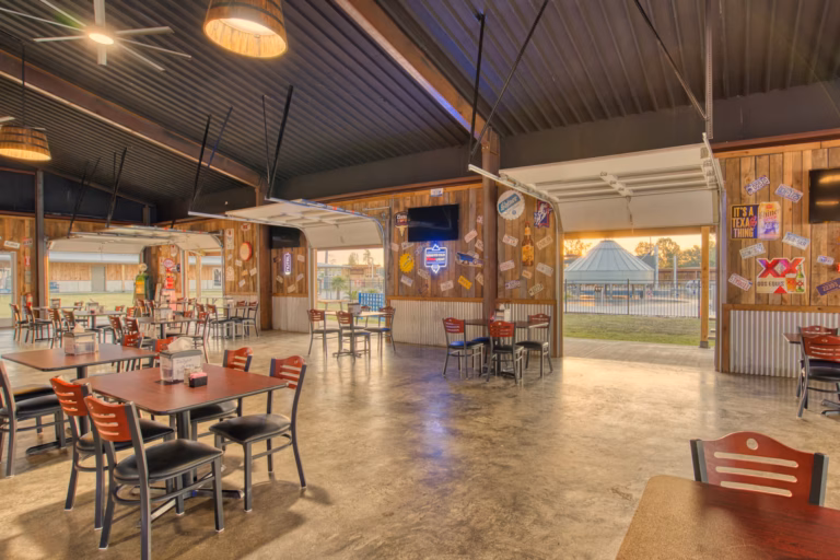 Open-air restaurant with wooden walls, large garage doors, various signs, and empty tables and chairs; outside view includes a pool area and trees at sunset.