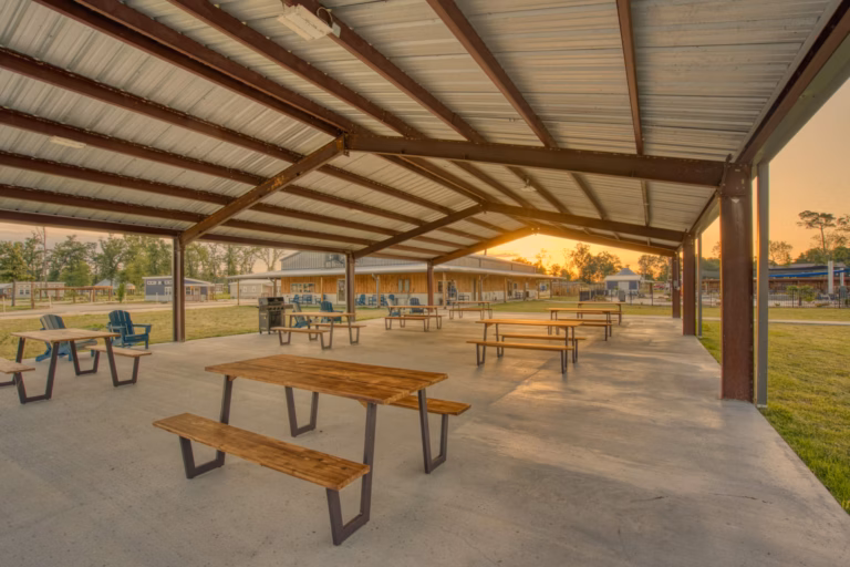 Open-air pavilion with a metal roof, several wooden picnic tables, and chairs on a concrete floor, with sunset light and grassy surroundings in the background.