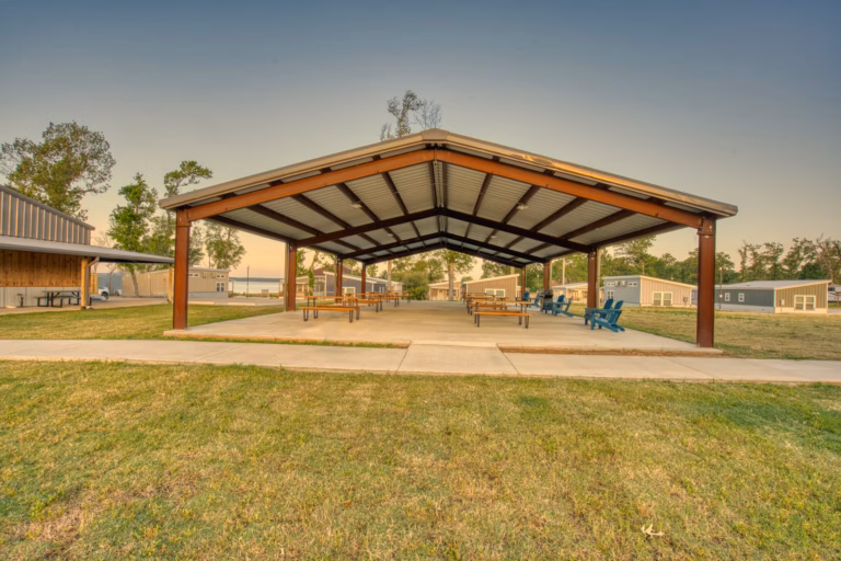A large open-sided metal pavilion with picnic tables and blue chairs on a concrete slab, surrounded by grass and nearby buildings under a clear sky.