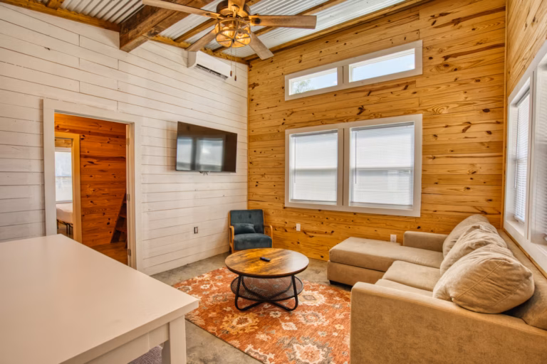 A small living room with wood-paneled walls, a beige sectional sofa, round coffee table, wall-mounted TV, armchair, and an orange patterned rug under a ceiling with exposed beams.