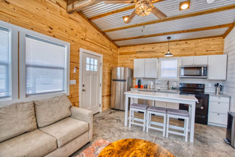 A small modern kitchen with white cabinets, stainless steel appliances, a breakfast bar with stools, a beige sofa, and wood-paneled walls and ceiling.