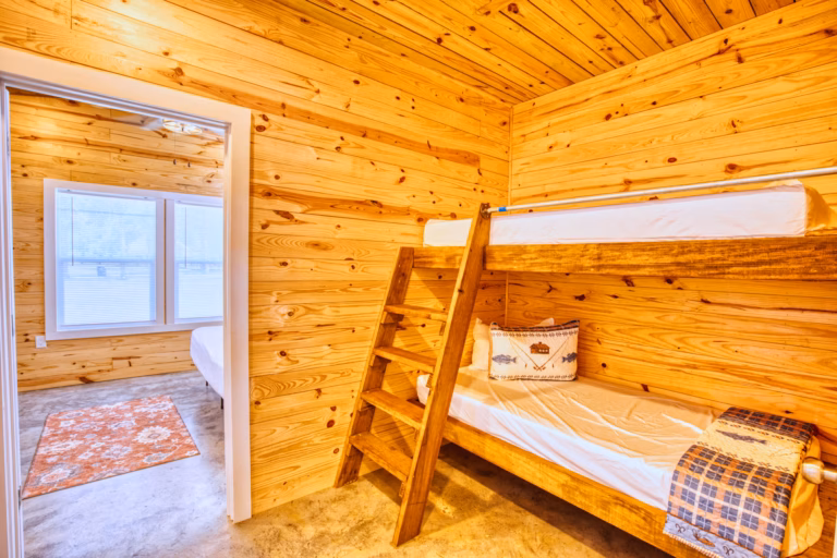 A wooden room with a bunk bed, ladder, fish-themed pillow, and a blue blanket; open doorway leads to a sunlit room with windows and an orange rug.