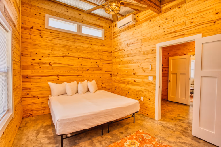 Bedroom with wood-paneled walls, a white bed with four pillows, concrete floor, ceiling beams, and a small orange rug near the bed.