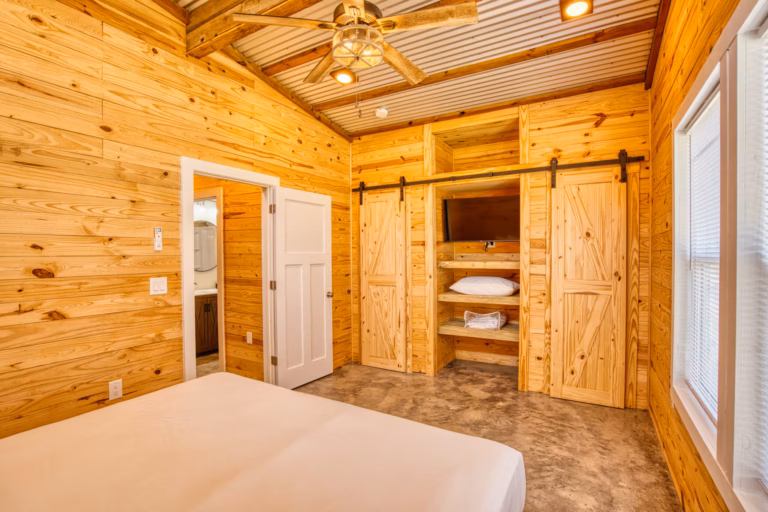 Bedroom with light wood paneling, a ceiling fan, a bed, an open closet with shelves, pillows, towels, and a wall-mounted TV. A door leads to a bathroom. Natural light from the window.