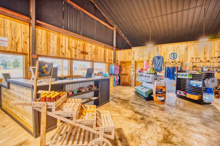 A farm store interior with wooden walls, a counter, shelves displaying shirts, hats, honey, jam, and other products under bright lighting.