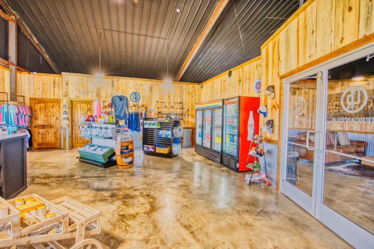 A rustic store interior with wooden walls, clothing racks, a refrigerated drinks section, and glass doors on the right.