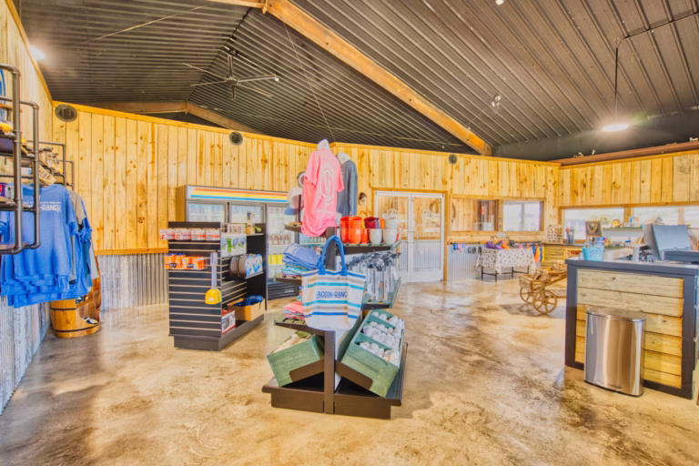 A rustic gift shop interior with wooden walls, merchandise on display racks, clothing, bags, and various items for sale, and a checkout counter on the right.