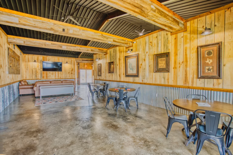 Rustic indoor seating area with wooden walls, metal chairs around round tables, framed art, a TV, and a sofa in the background on a polished concrete floor.
