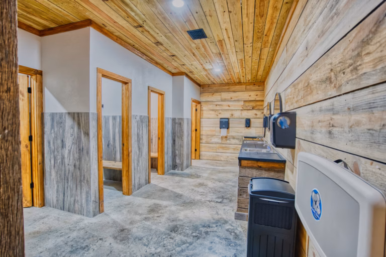Modern public restroom with wooden walls and ceiling, stalls on the left, sinks and mirrors on the right, and a baby changing station in the foreground.