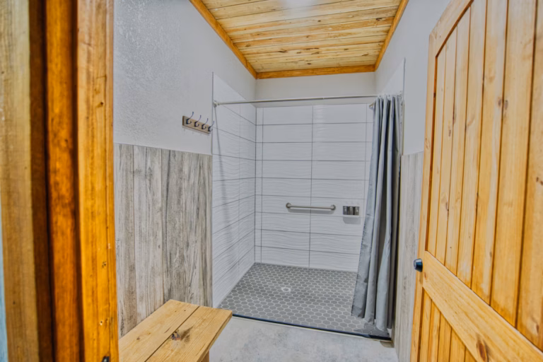 Accessible shower with grab bar, gray curtain, tiled walls, and wooden accents, including a door, bench, and ceiling. Four hooks are mounted on the left wall.