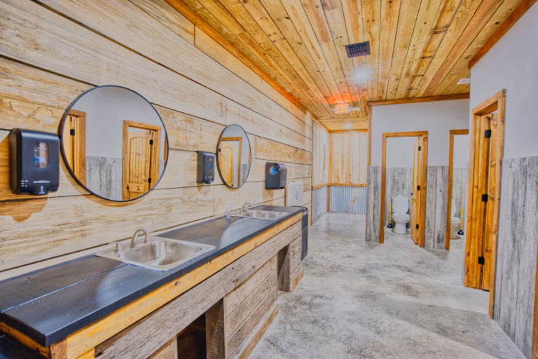 A rustic public restroom with wooden walls and ceiling, three sinks with mirrors, soap dispensers, and stalls with toilets in the background.