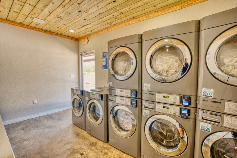 A small laundry room with six stainless steel front-loading washers and dryers stacked against the wall, featuring concrete floors and a wooden ceiling.