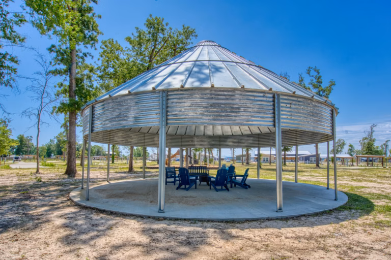 A round metal pavilion with open sides shelters a circle of blue chairs and a table, set on a concrete pad in a park with trees and clear sky.