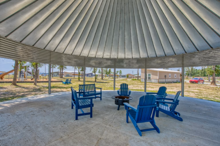 Six blue Adirondack chairs surround a fire pit under a large, open-sided metal pavilion, with buildings and trees visible outside.