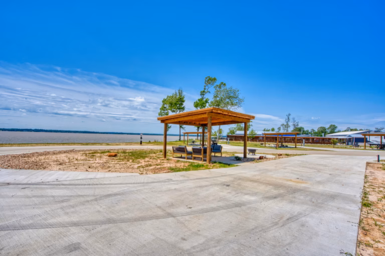 A wooden gazebo with outdoor seating is set on a concrete lot near a sandy, open landscape under a clear blue sky.