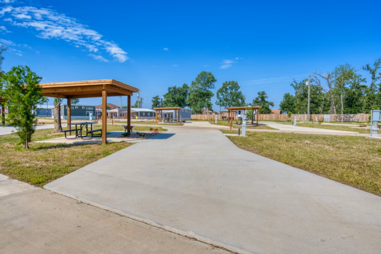 A paved RV site with a wooden picnic shelter and table, surrounded by grass and trees, under a clear blue sky. Multiple similar sites are visible in the background.