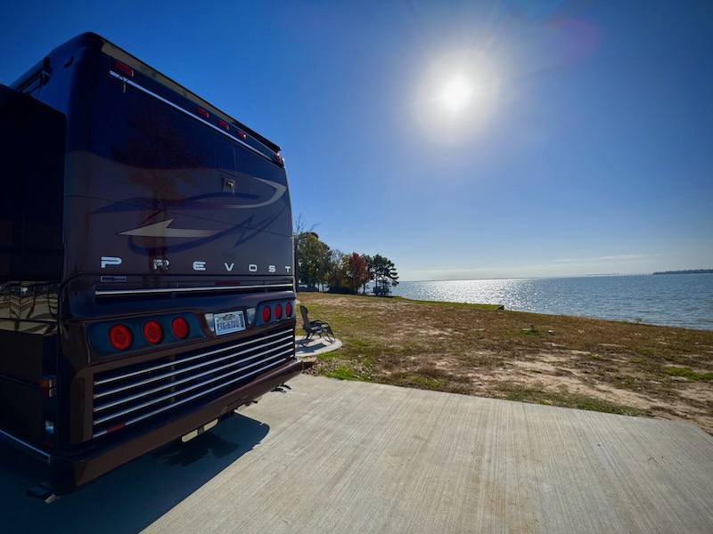 A large recreational vehicle is parked on a concrete pad near a grassy shoreline, with the sun shining over a calm body of water.