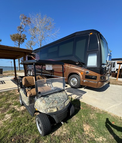 A camouflage-patterned golf cart is parked in front of a large brown and black tour bus on a sunny day, with a lake and trees in the background.