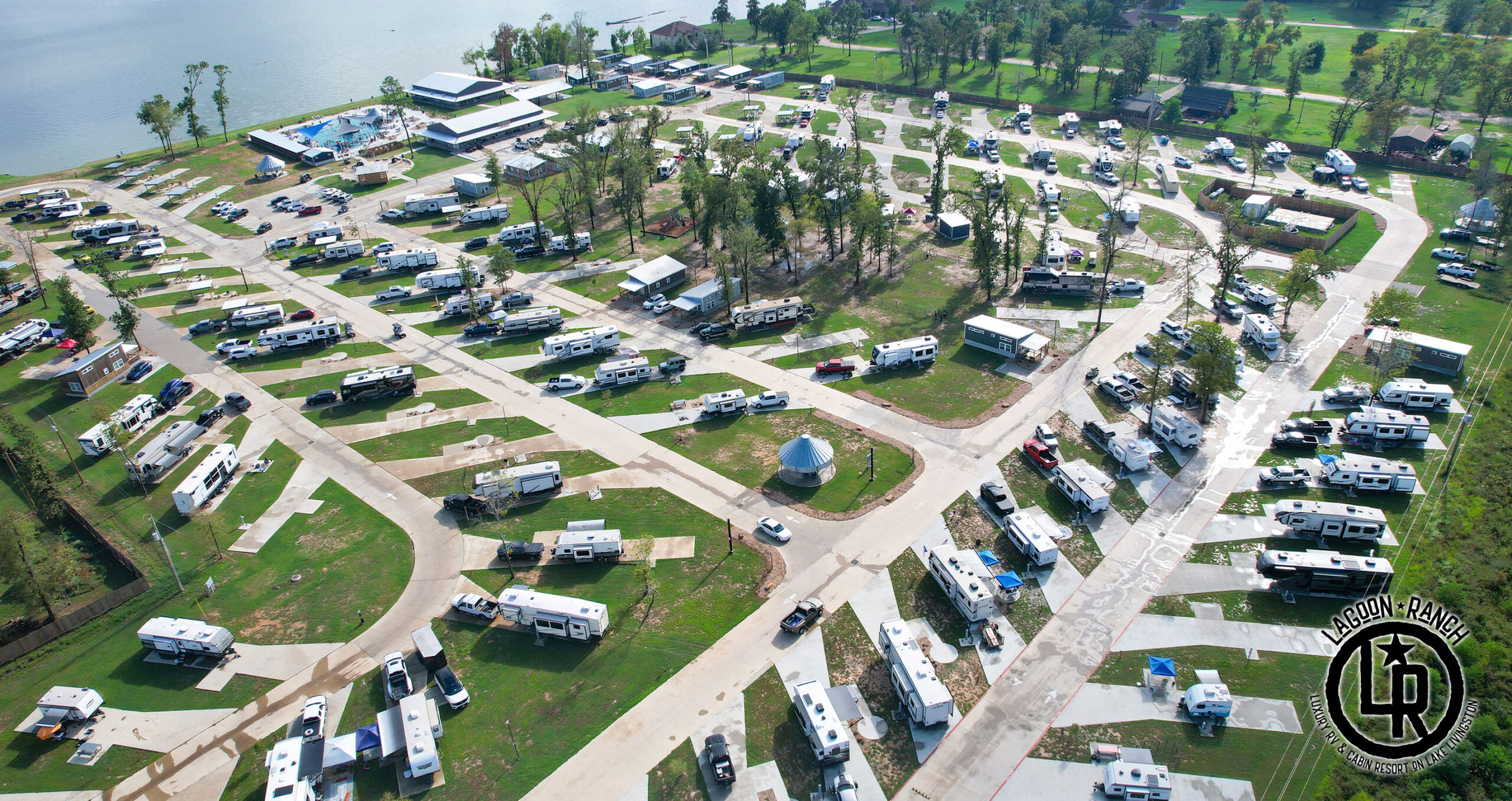 Aerial view of an RV park with numerous trailers parked in organized rows, surrounded by trees, green grass, and nearby buildings.