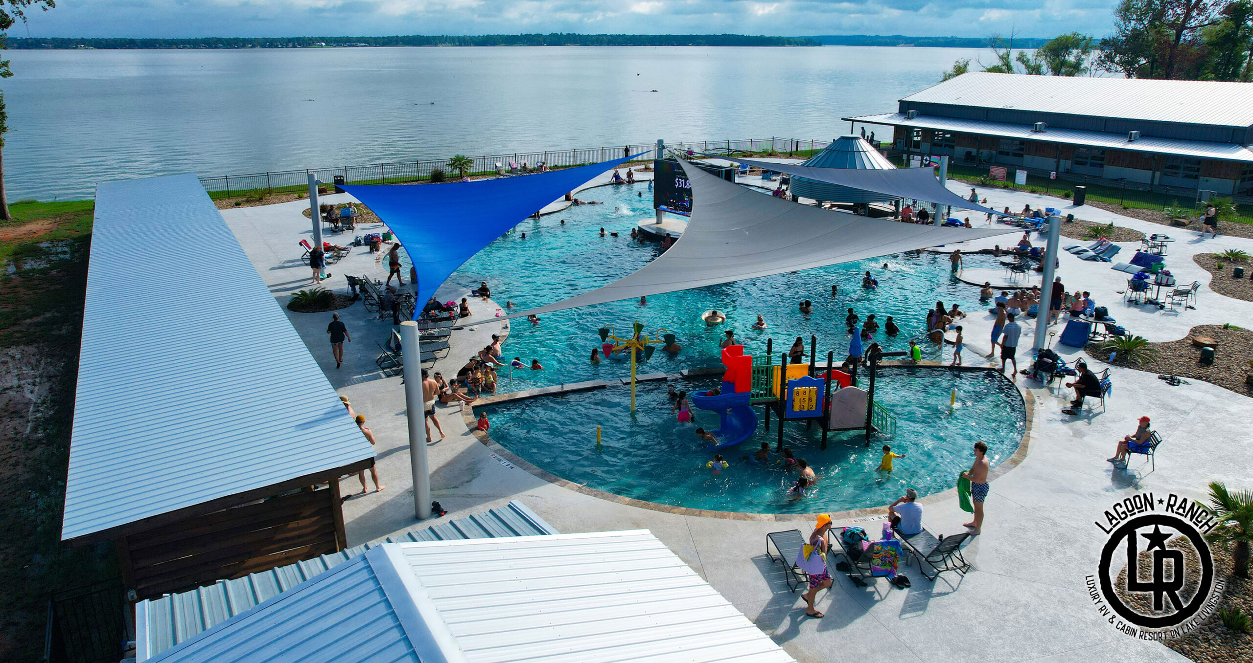 An outdoor public pool by a lake features people swimming, relaxing, and playing; shaded areas, lounge chairs, and a building are visible nearby, with the Larson Ranch logo in the corner.