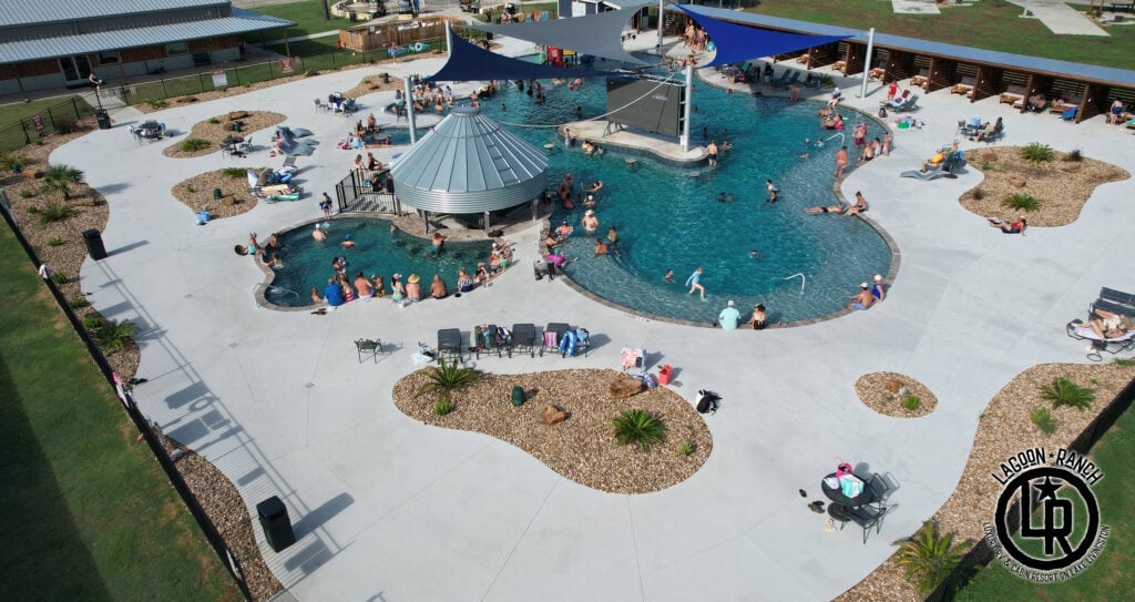 Aerial view of a large outdoor pool with people swimming and lounging, surrounded by chairs, shaded areas, and landscaped walkways.