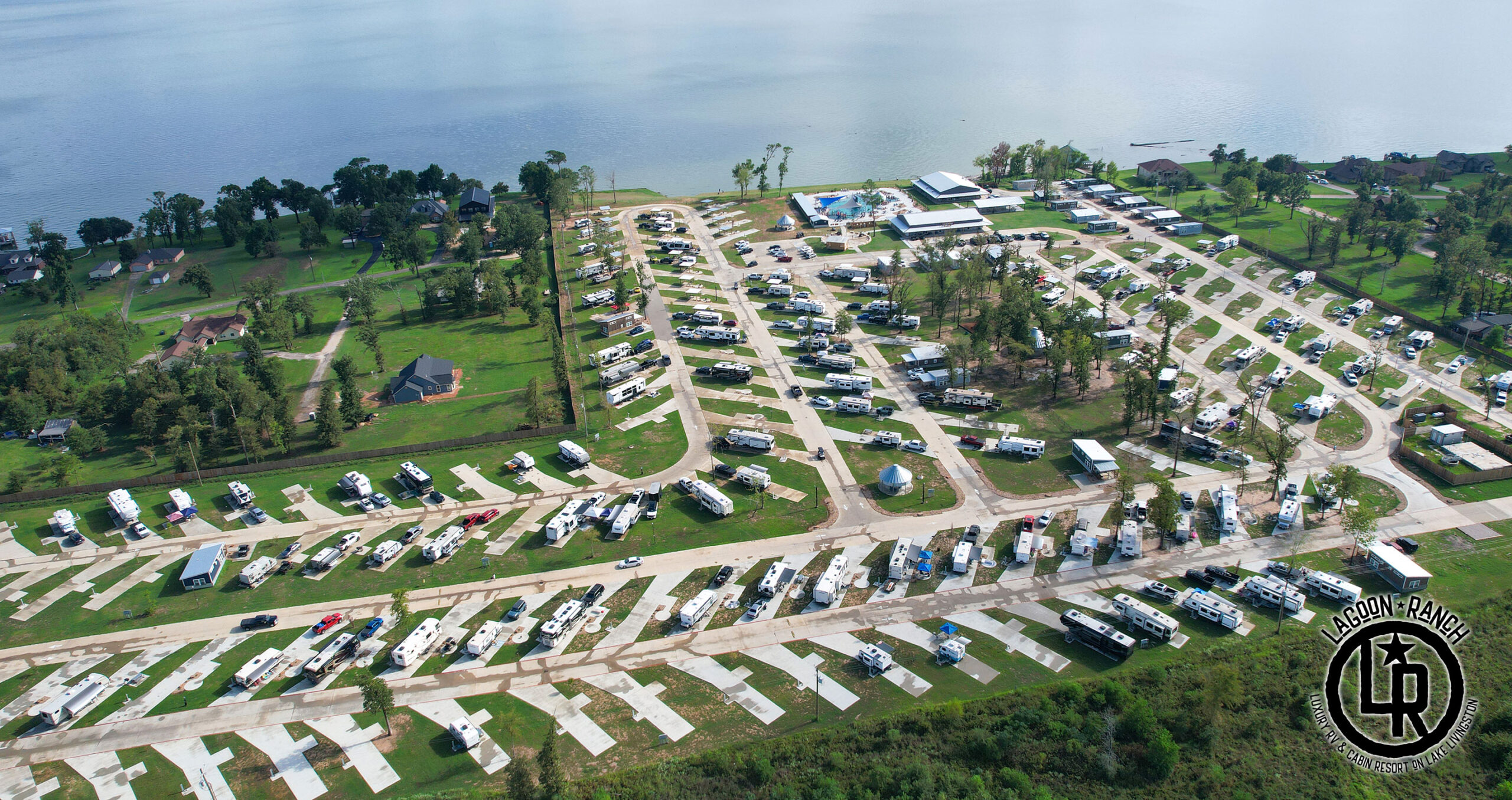 Aerial view of an RV park with numerous parked RVs, adjacent to a lake, surrounded by trees and houses, with a central building and pool visible.