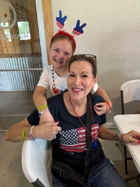 A woman and a girl, both wearing patriotic clothing and accessories, smile while sitting at a table indoors.