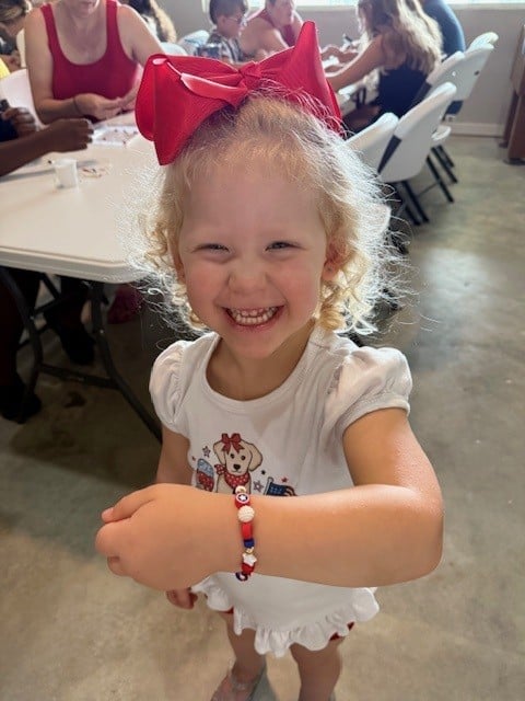 A young girl with a big red bow smiles and shows off a beaded bracelet at a table with other children in the background.