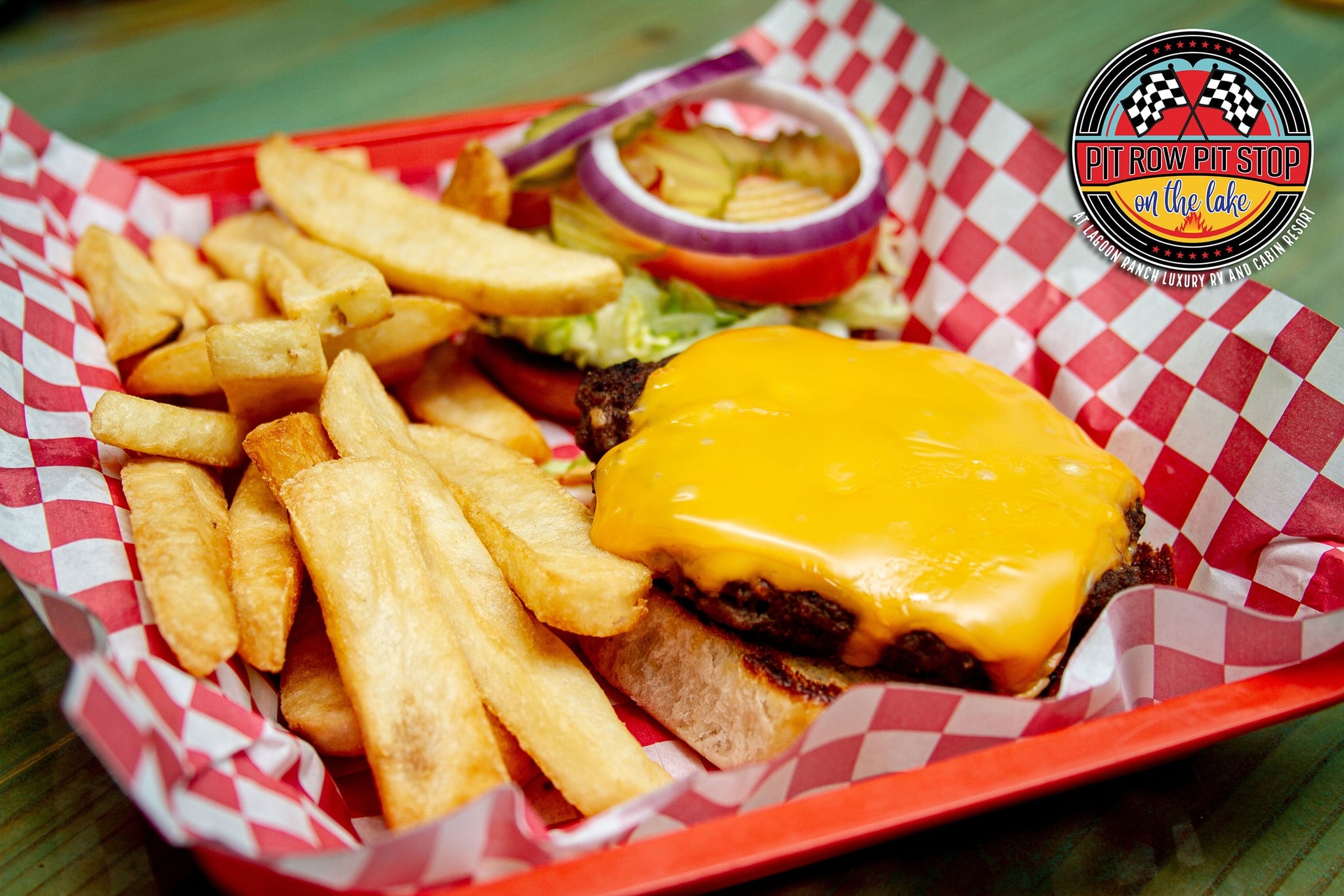A cheeseburger with lettuce, tomato, pickles, and onion served with thick-cut fries on a red tray with a Pit Row Pit Stop logo in the corner.