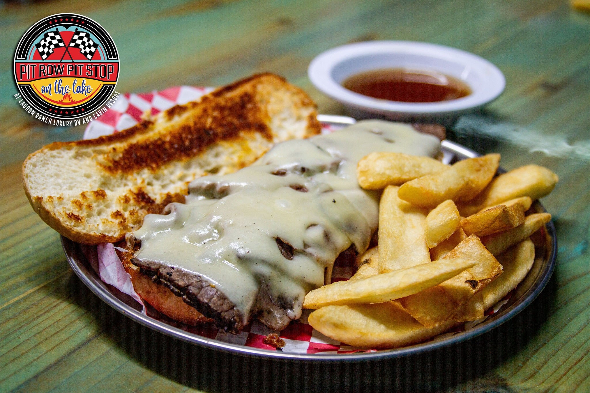 A sandwich with melted cheese and beef, served with French fries and a side of dipping sauce on a plate; a "Pit Row Pit Stop" logo is visible in the image.