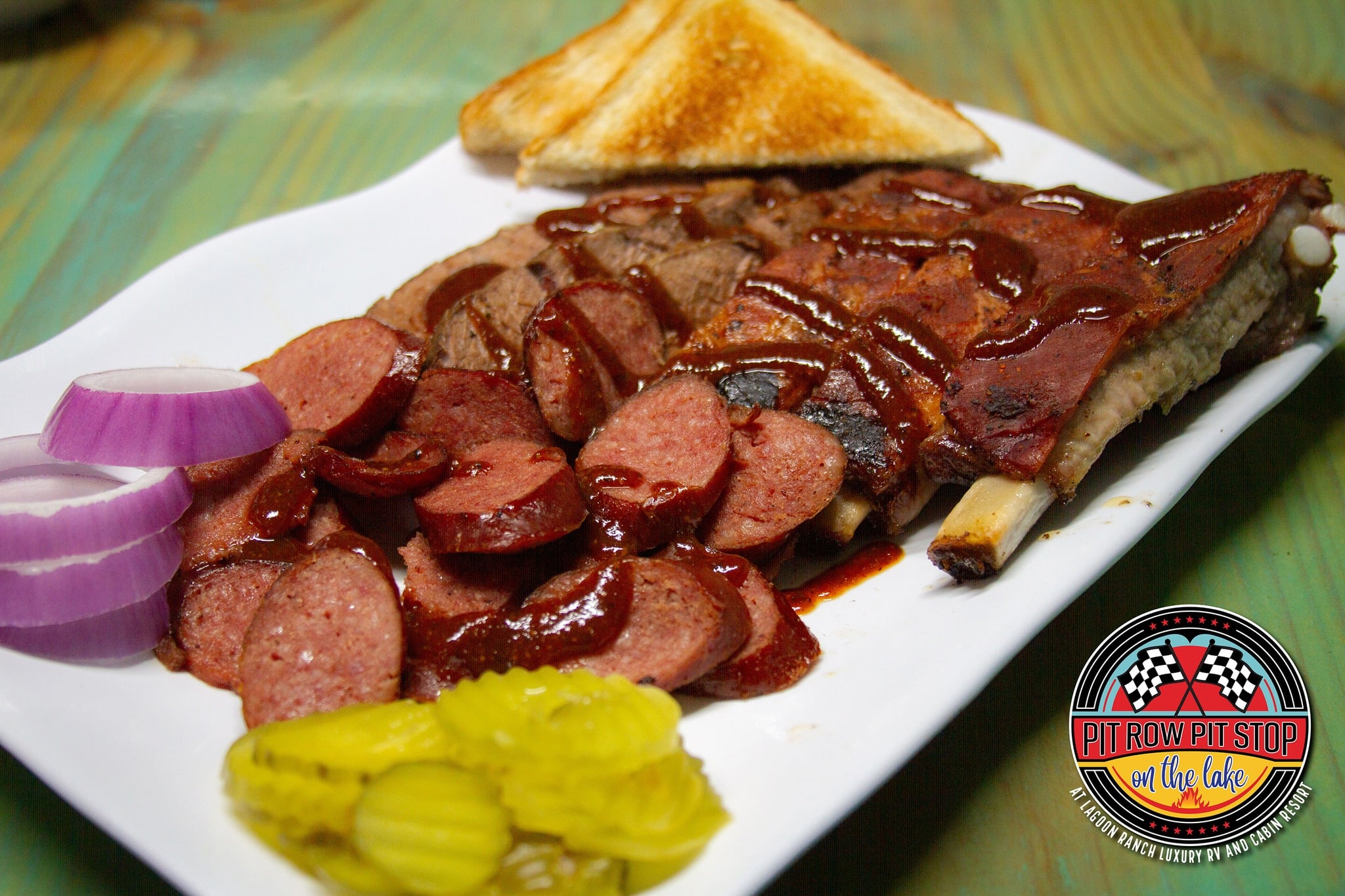 A plate with barbecued ribs, sliced sausage, sliced red onion, pickles, and two pieces of toast. A logo reading "Pit Row Pit Stop on the Lake" is visible in the corner.