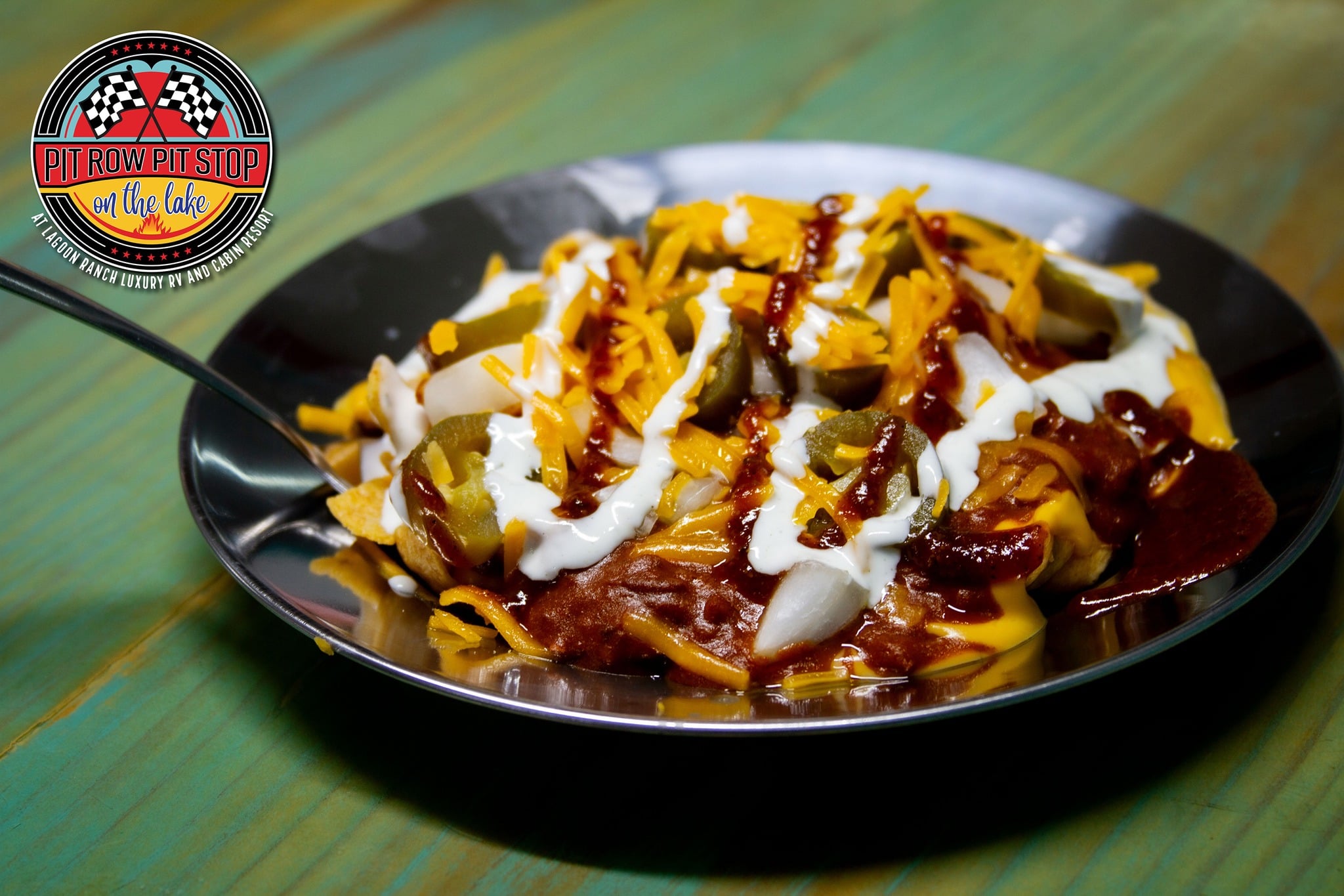 A plate of loaded nachos topped with melted cheese, sour cream, jalapeños, chopped onions, and barbecue sauce sits on a green wooden table. Logo in upper left corner reads "Pit Row Pit Stop.