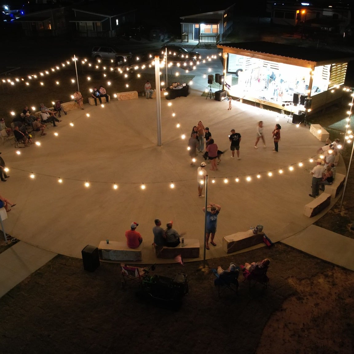 A group of people gather under string lights in an outdoor circular concrete area at night, watching a band perform on a lit stage.