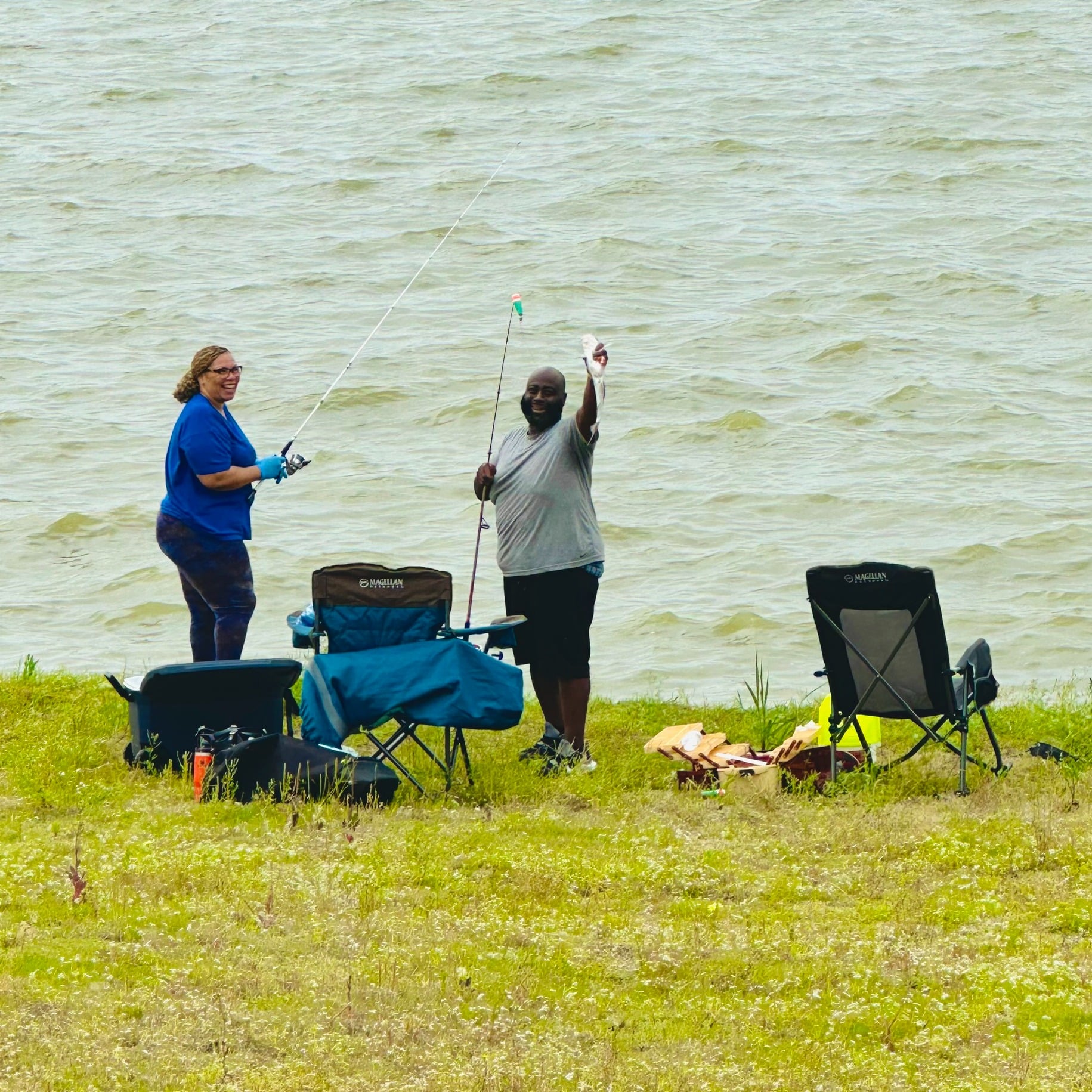 Two people stand by a lakeshore holding fishing rods, with chairs and gear set up nearby on the grass. One person appears to be showing a catch.