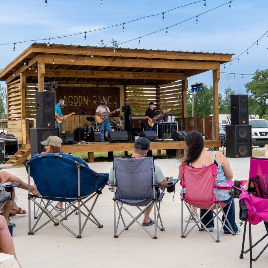People sit in folding chairs watching a live band perform on an outdoor wooden stage under string lights during the day.