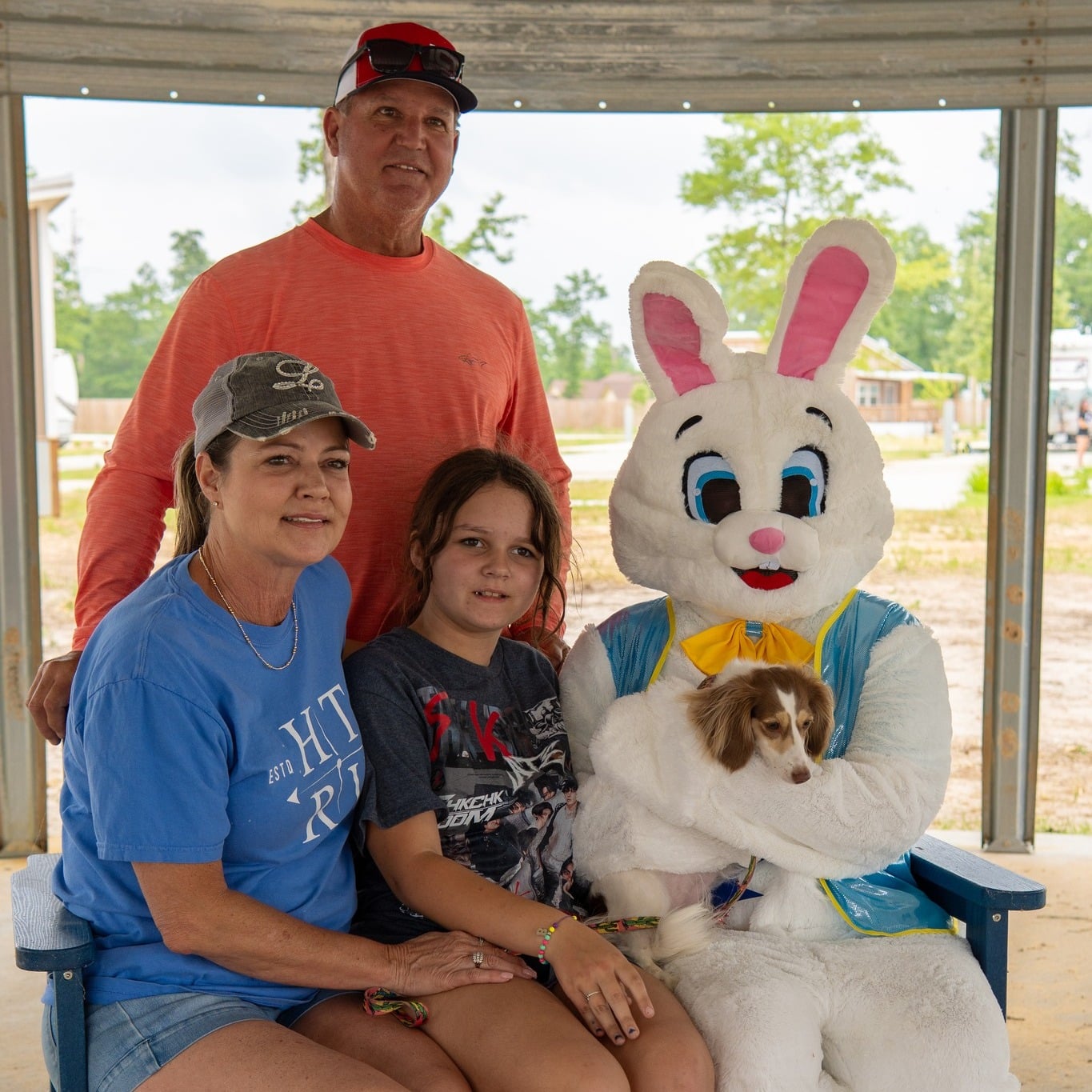 A family of three poses with a person in an Easter Bunny costume holding a small dog, all sitting on a bench inside a covered outdoor area.