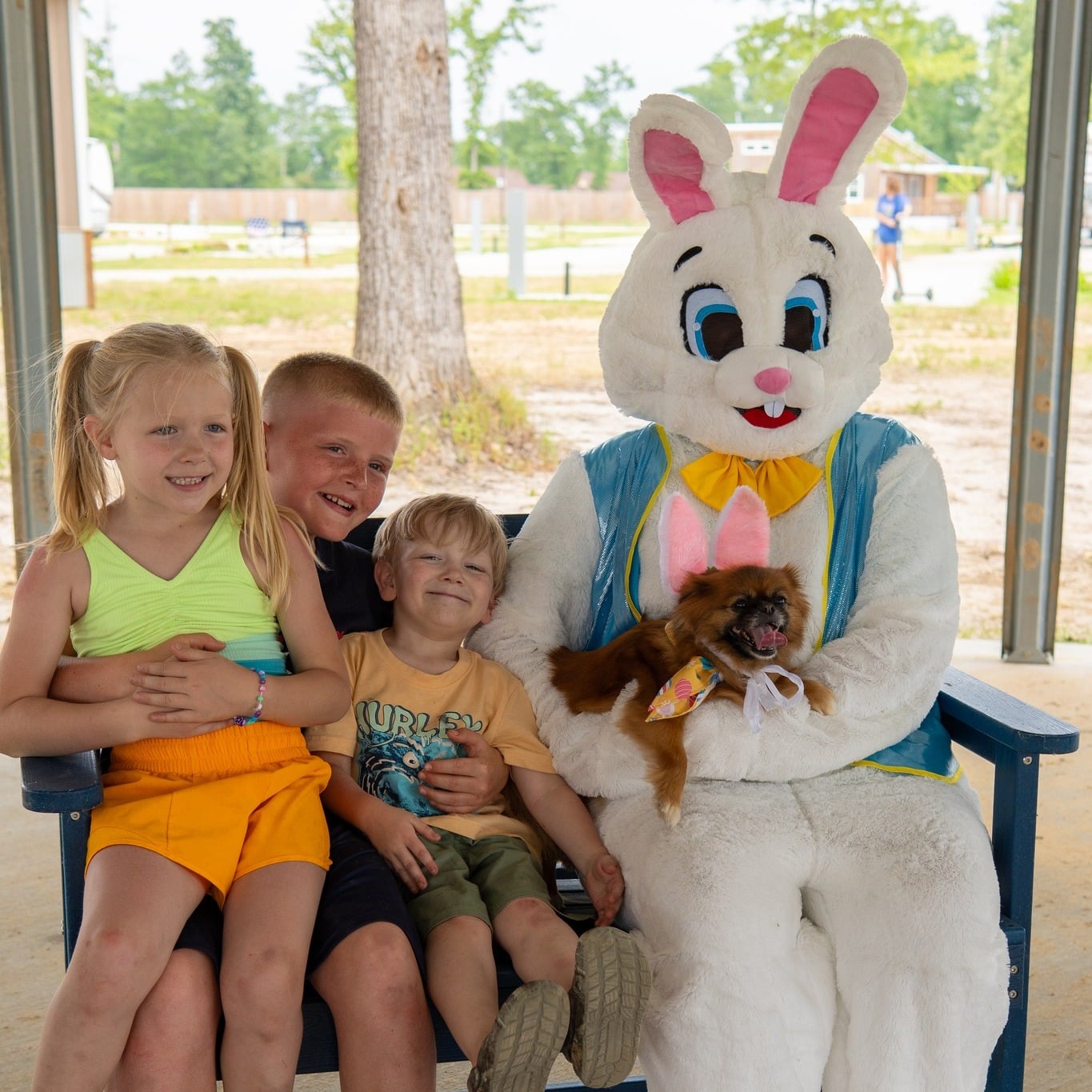 Three children and a small dog sit on a bench with a person in an Easter Bunny costume under a shelter, with trees and a field in the background.