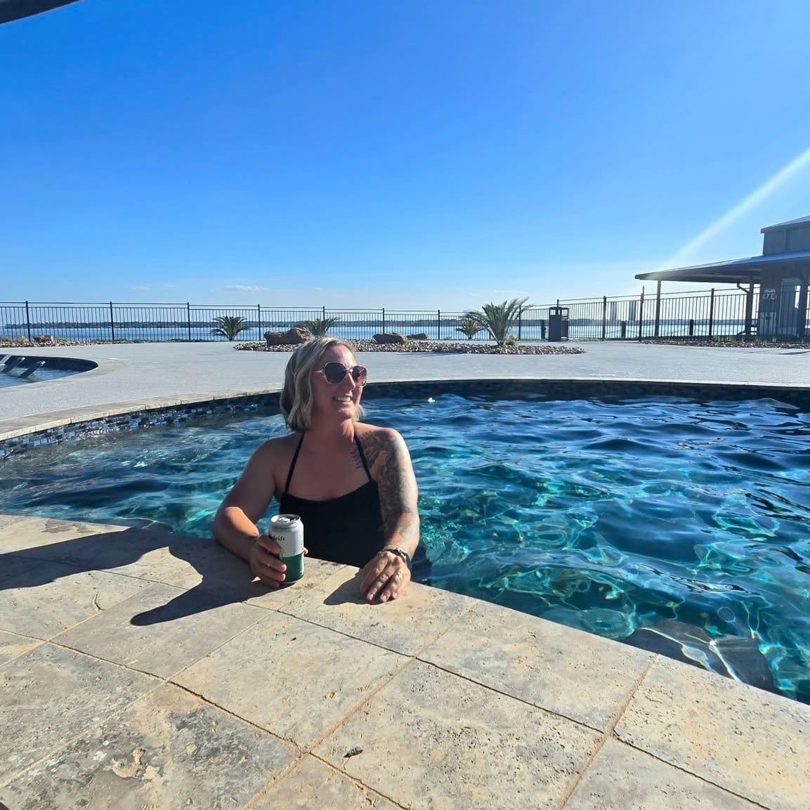 A woman with sunglasses and a can of drink relaxes in a swimming pool on a sunny day, with a clear blue sky and a fence in the background.