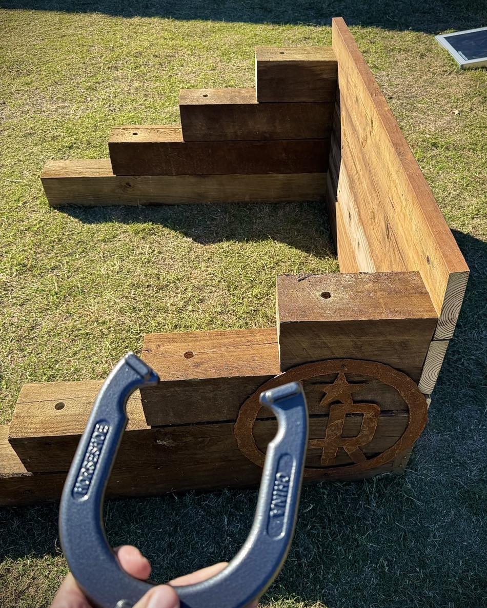 A person holds a horseshoe in front of a wooden horseshoe pit with tiers, set up on grass in an outdoor area.