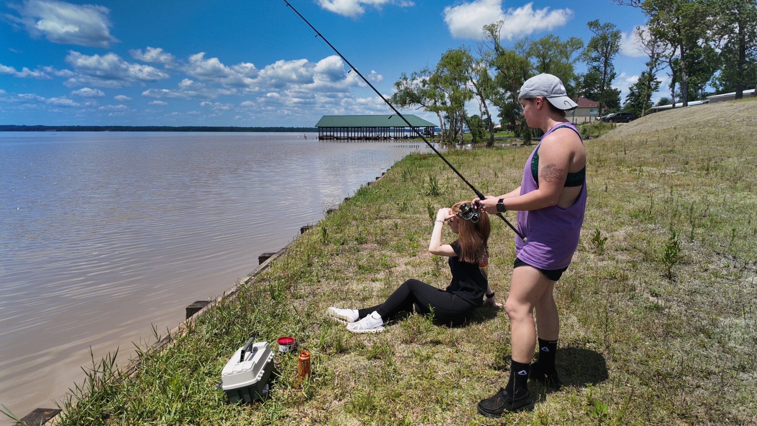 Two people fishing by a riverbank; one is standing and holding a fishing rod, while the other sits on the grass beside a tackle box and drinks from a bottle.
